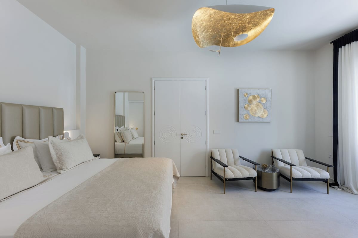 Une chambre d’hôtel moderne comprenant un lit, deux fauteuils, une table basse ronde, un grand miroir et un luminaire au plafond au design original.