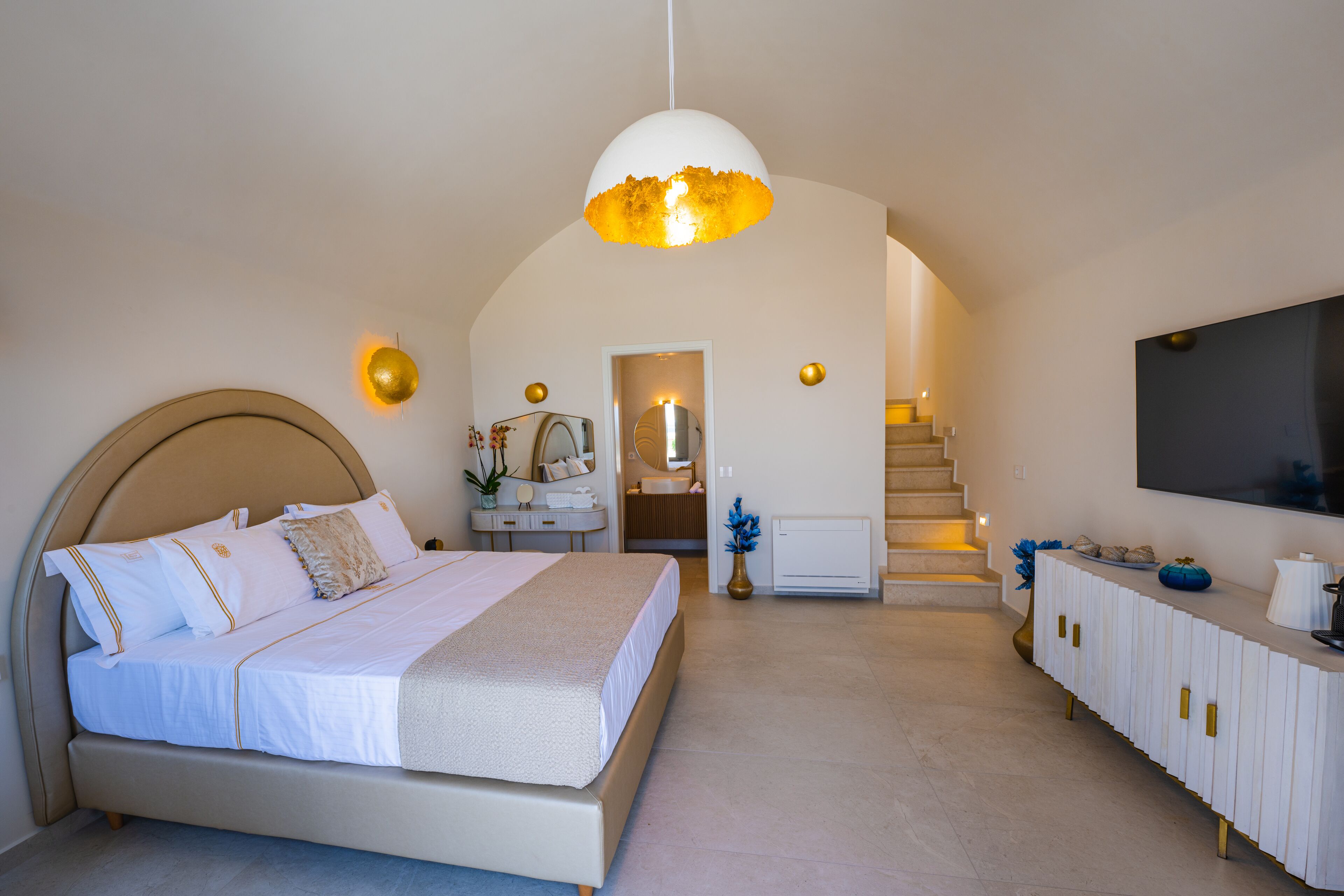 A bedroom with a large bed, a vanity with a mirror, a television mounted on the wall, and a staircase leading to an upper level.