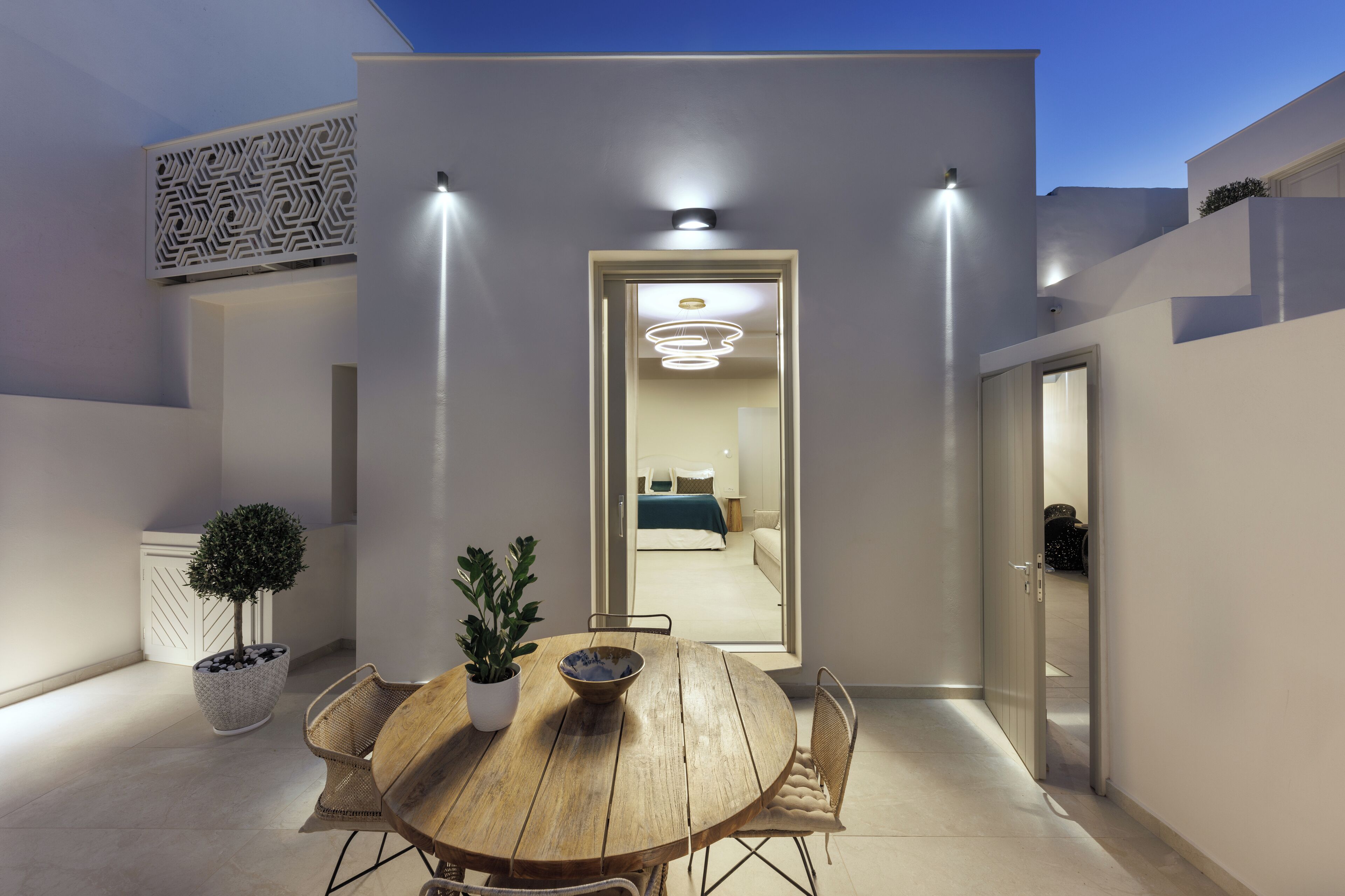 A modern outdoor dining area with a wooden table and chairs, potted plants, and a view into a bedroom through an open door.