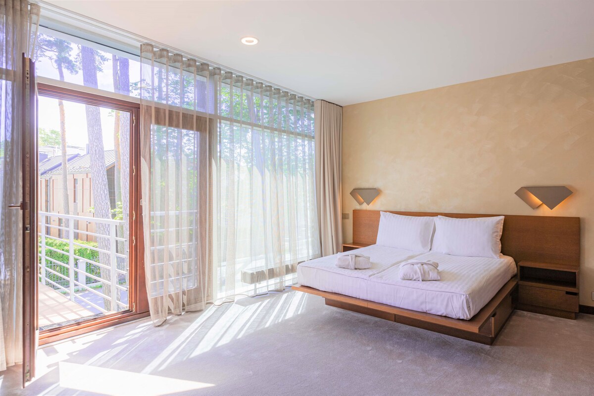 Un dormitorio con cama, mesita de noche, ventana con cortinas y vistas al exterior.