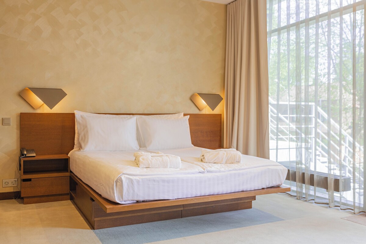 Habitación de hotel con una cama grande, dos lámparas de noche, una mesita de noche y vistas a un balcón con barandilla.
