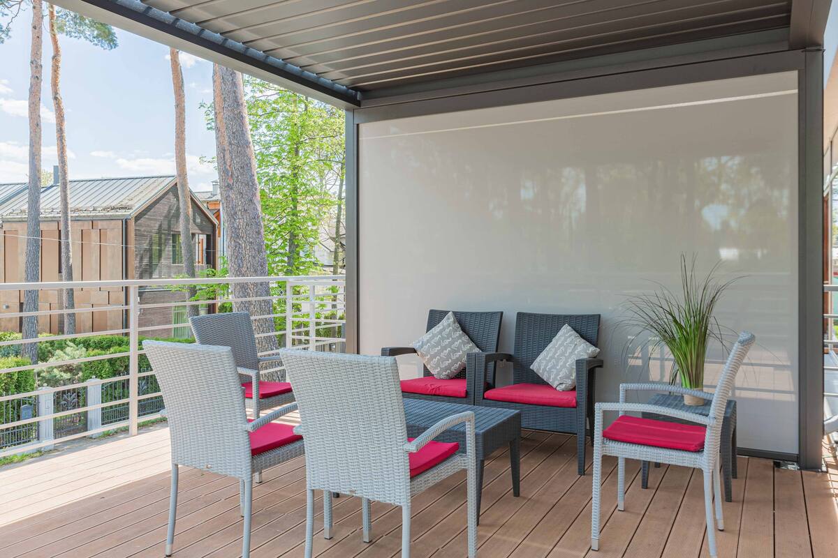 A covered outdoor seating area with wicker chairs and red cushions, a potted plant, and a view of a wooden deck and trees.