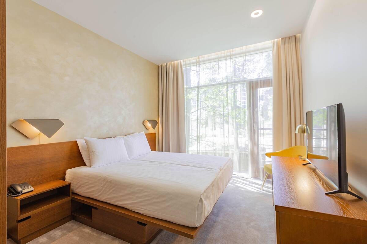 A hotel room with a large bed, bedside tables, a desk with a chair, and a TV.