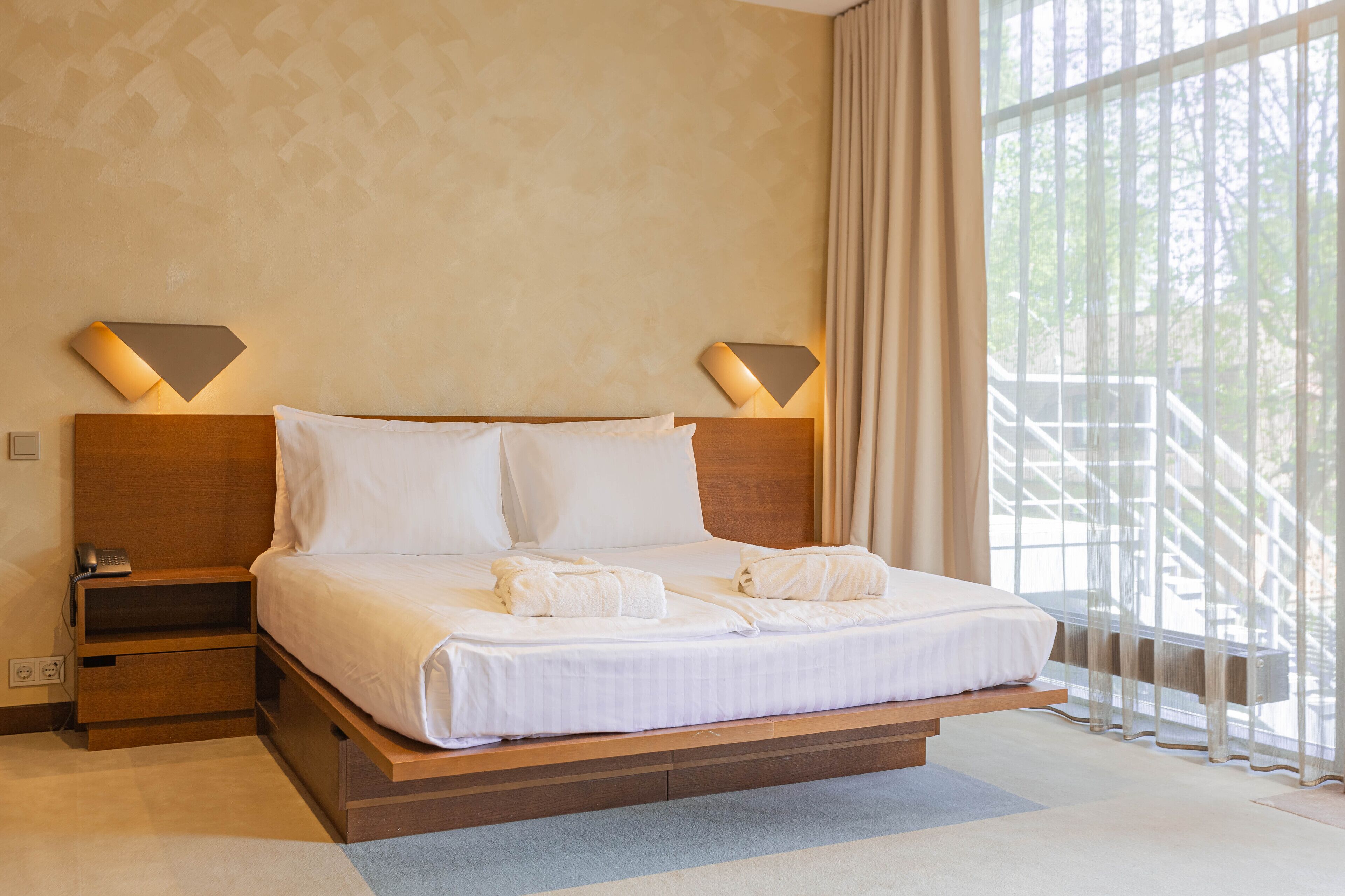 A hotel room with a large bed, two bedside lamps, a nightstand, and a view of a balcony with a railing.