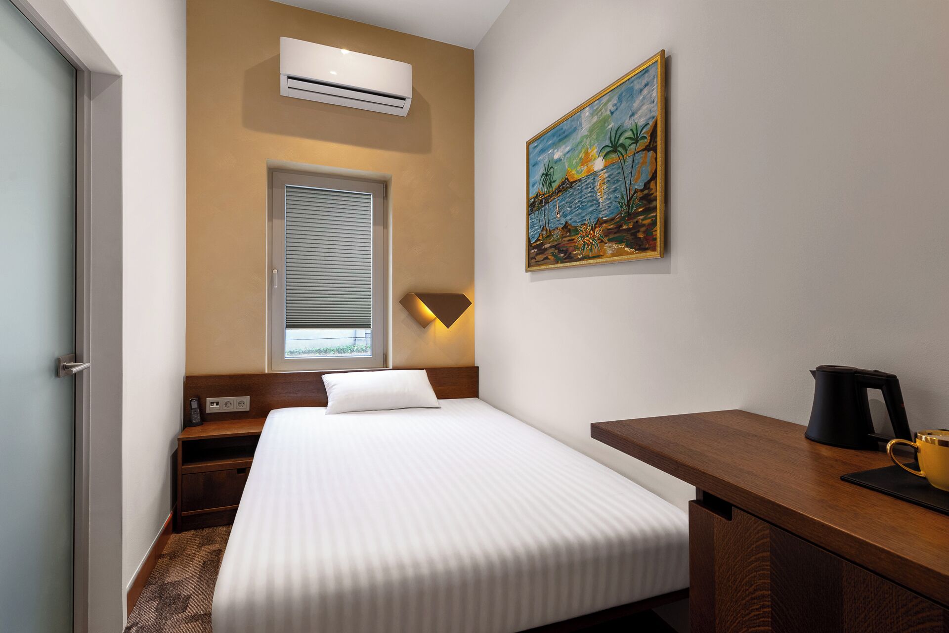 A hotel room with a bed, bedside table, lamp, and a painting on the wall.