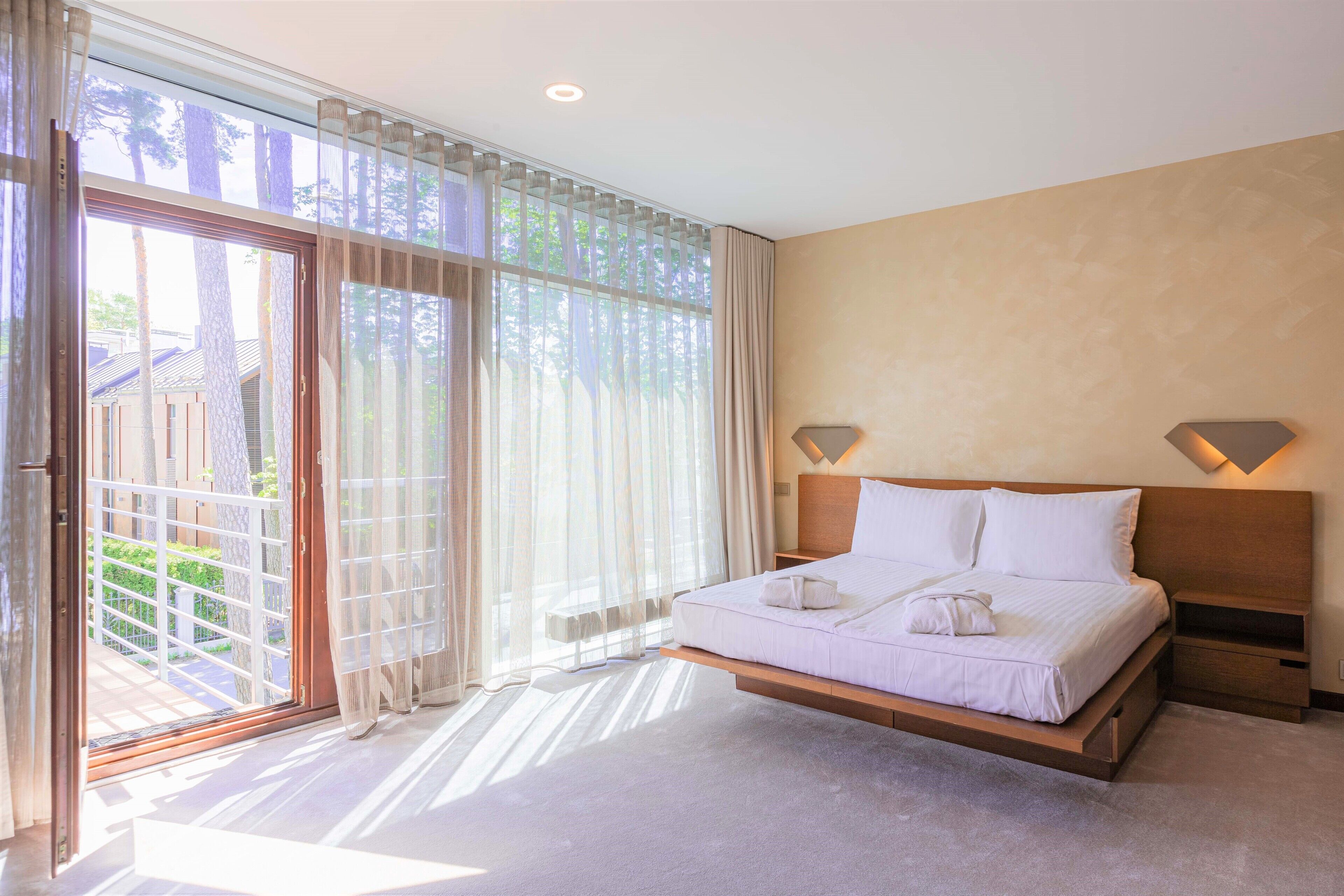 A bedroom with a bed, a nightstand, a window with curtains, and a view of the outside.