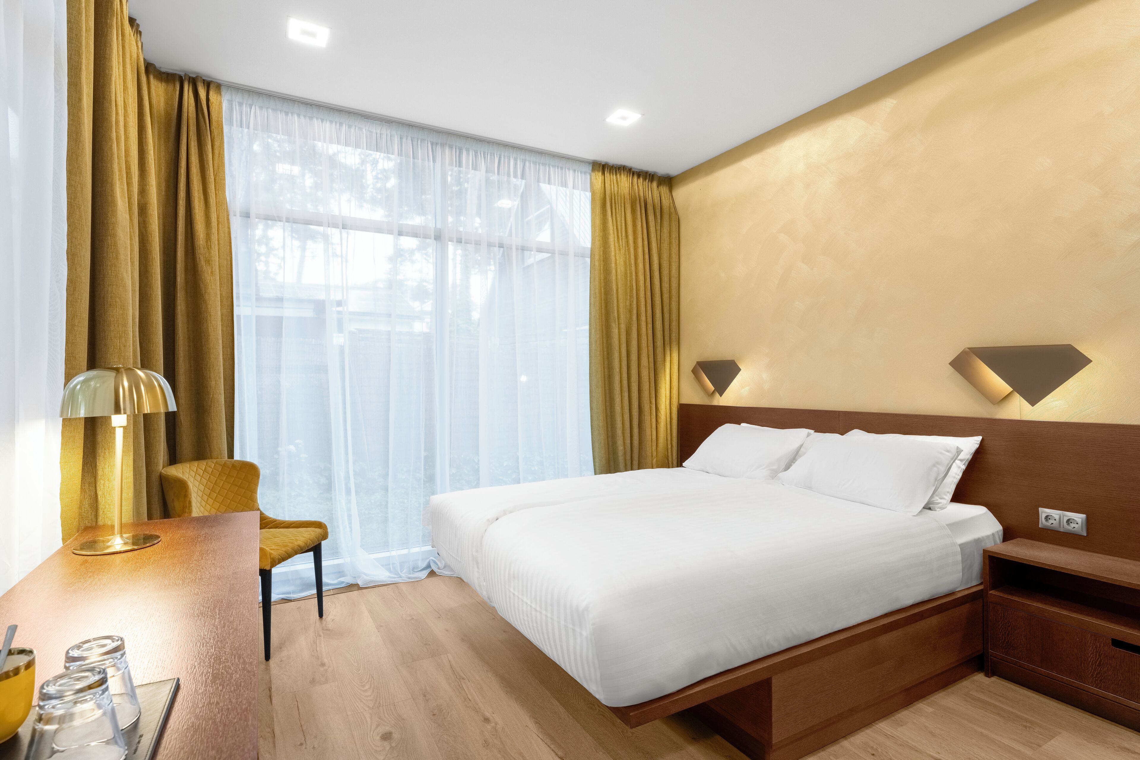 Habitación de hotel con una cama grande, un escritorio con lámpara, una silla y un ventanal con cortinas.