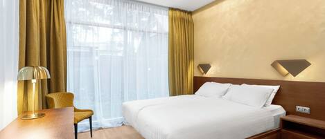 Habitación de hotel con una cama grande, un escritorio con lámpara, una silla y un ventanal con cortinas.