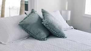 En pænt redt seng med et hvidt quiltet sengetæppe og to puder, hvoraf den ene har et struktureret mønster og den anden en detalje med en kvast.