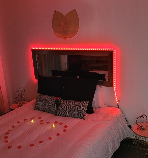 Una cama con espejo e iluminación LED, una decoración con forma de corazón sobre el espejo, y pétalos de rosa sobre la cama.