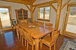 A wooden dining table with eight chairs, a cabinet with glass doors, and large windows offering a view of a grassy field and mountains.