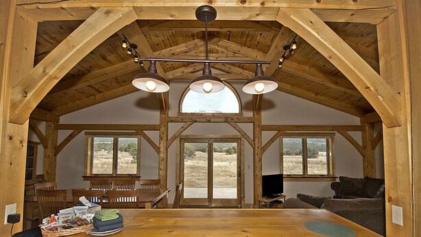 A rustic interior with a wooden dining table, benches, and a sofa. There are three windows providing views of the outdoors. - Whispering Hills Retreat (Saguache County)