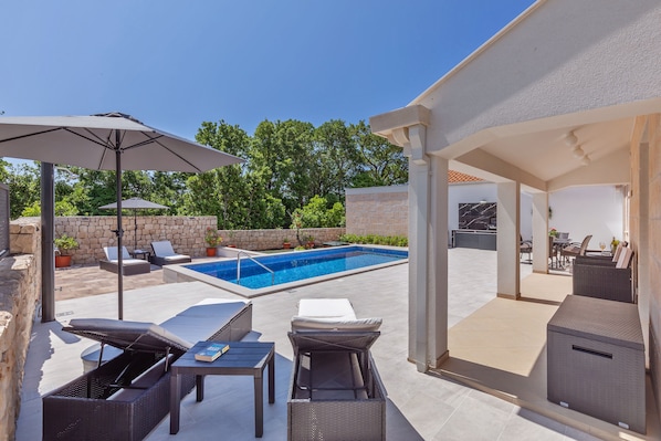 Uma área à beira da piscina com espreguiçadeiras, uma mesa e uma churrasqueira.