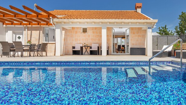 A swimming pool with clear blue tiles, a covered patio area with outdoor furniture, and a building with a tiled roof.