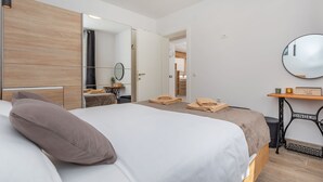 A modern hotel room with a large bed, a wooden headboard, a round mirror, a small table with a lamp, and a view into the hallway.
