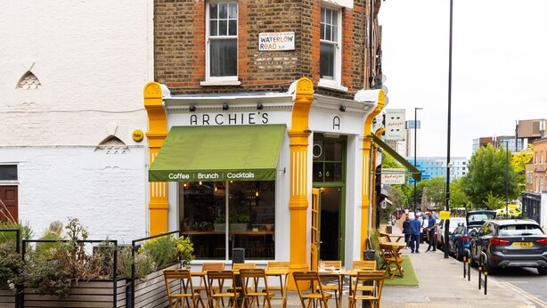 A small, yellow and white building with a green awning labeled "ARCHIE'S" offering coffee, brunch, and cocktails. - Stylish one bedroom apartment in Greater London (London)