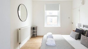 A bedroom with a large bed, a radiator, a window with blinds, a mirror, and a small shelf. - Stylish one bedroom apartment in Greater London (London)