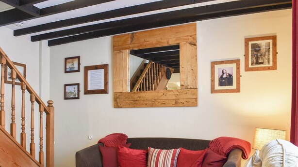 Un salón acogedor con una escalera de madera, un sofá gris oscuro con cojines rojos y a rayas, una mesa de centro de madera y un espejo en la pared.