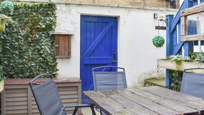 Un área de estar al aire libre de estilo rústico con una mesa y sillas de madera, una puerta azul y un muro de piedra con hiedra.