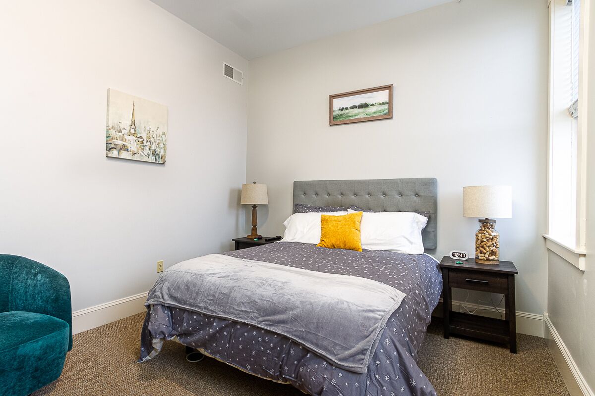 A bedroom with a bed, two nightstands, a lamp, a chair, and two wall art pieces.