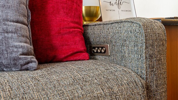 Un canapé gris et rouge à texture marquée, doté d’un panneau de commande visible, une lampe dorée et un panneau encadré sur lequel figurent les mots « wifi » et « mot de passe », le tout posé sur une surface en bois.