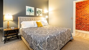Une chambre à coucher avec une tête de lit capitonnée, une couvre-lit à motifs, une table de chevet avec une lampe et des œuvres d’art accrochées au mur.