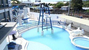 Una piscina con un tobogán espiral azul y un pequeño puente, rodeada de sillones blancos y un edificio blanco.