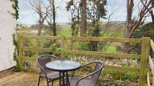 Eine Terrasse im Freien mit einem runden Glastisch und zwei Korbstühlen, umgeben von einem Holzzaun und Pflanzen.