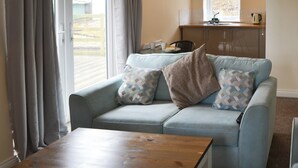 Ein Wohnzimmer mit einem hellblauen Sofa, einem Holztisch, einer Küchenzeile mit Spüle und Schränken sowie einem Fenster mit Vorhängen.