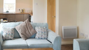 Ein Wohnzimmer mit einem hellblauen Sofa, zwei gemusterten Kissen, einem Holztisch und einem Küchenbereich im Hintergrund, der ein Fenster, ein Spülbecken und Geräte zeigt.