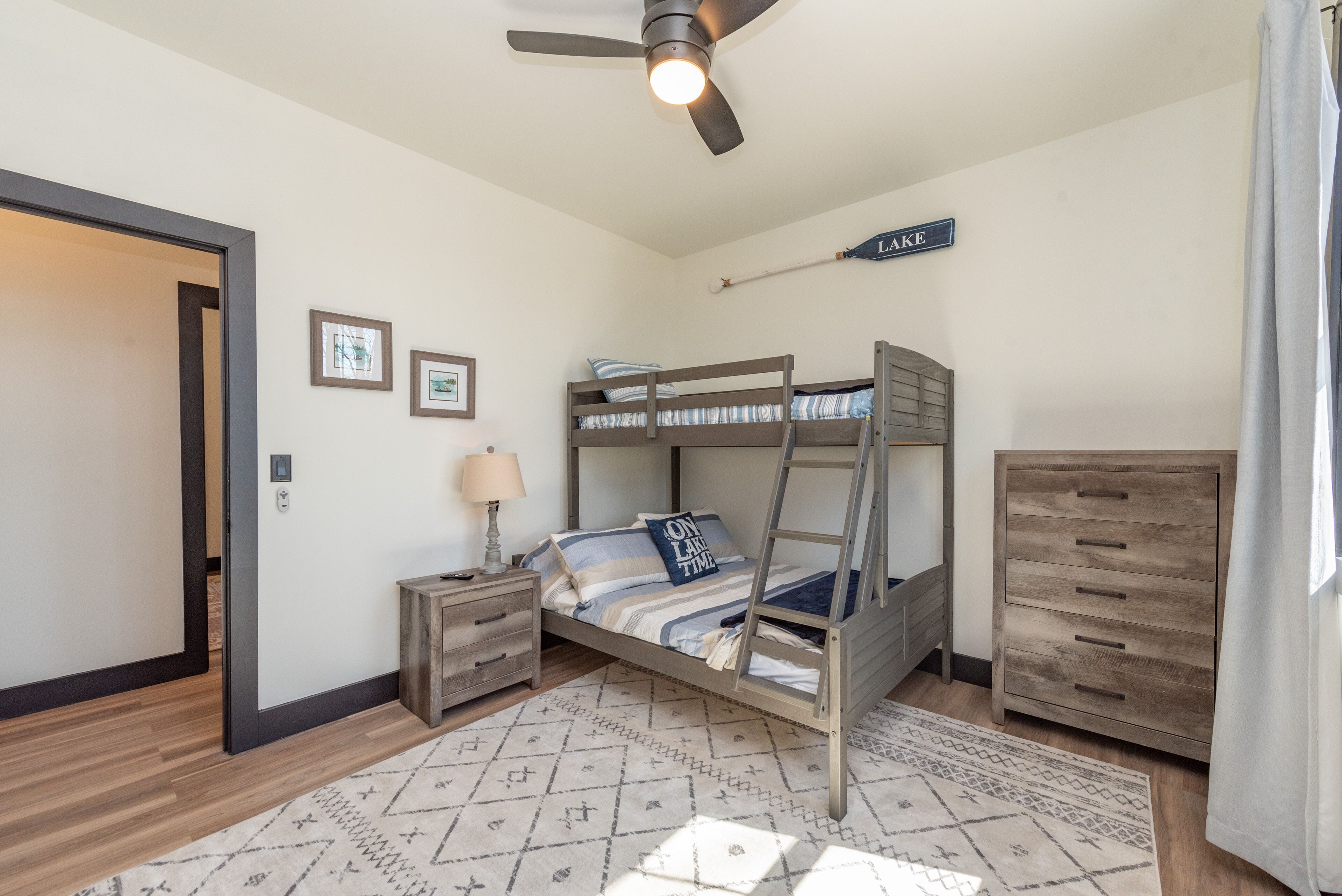 A bedroom with a bunk bed, a desk with a lamp, a dresser, a ceiling fan, and a sign on the wall.