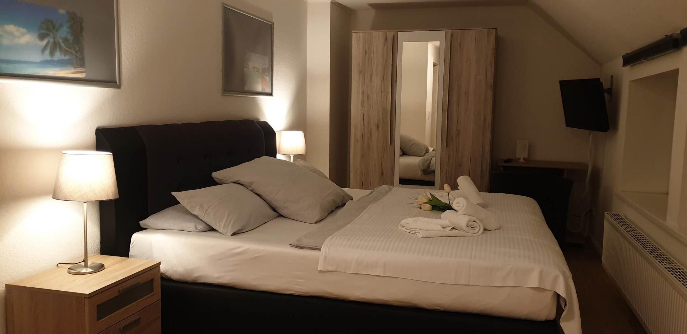 Ein ordentlich bezogenes Bett mit Kissen, ein Nachttisch mit Lampe, ein Spiegel und ein an der Wand befestigter Fernseher.