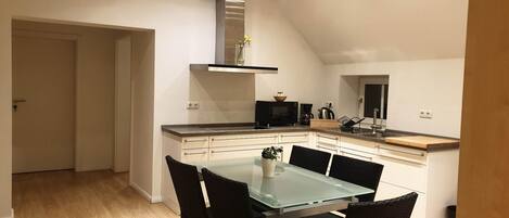 A modern kitchen with a dining area, featuring a glass table, black chairs, a stainless steel range hood, and a microwave.