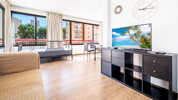 Een moderne woonkamer met een bank, een televisie, een bureau en een balkon met uitzicht op gebouwen en bomen.