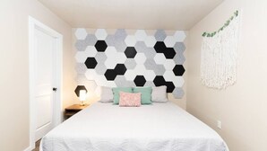 A bedroom with a bed, a wall-mounted geometric pattern, a macrame wall hanging, and a bedside lamp.