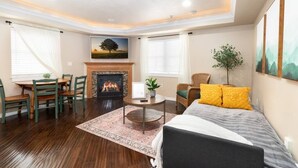 A living room with a fireplace, a dining area with wooden chairs, a bed with a grey and white bedspread, and a large window with a view of a tree.
