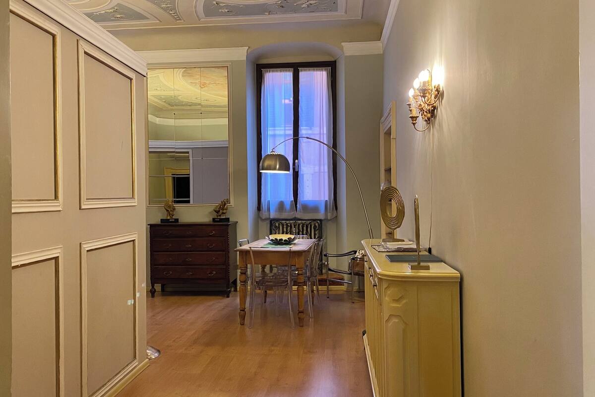 A hallway with a wooden floor, a dining table with chairs, a mirror, and a lamp.