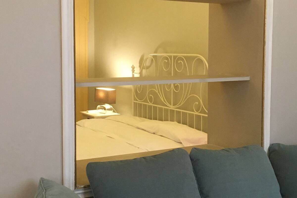 A room with a bed, a bedside table with a lamp, and a window with a view of a building.