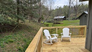 Um deck de madeira com duas cadeiras Adirondack e uma pequena mesa, com vista para uma área gramada com árvores e um prédio ao longe.