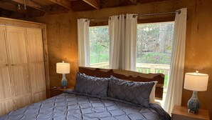 Quarto com uma cama grande, duas mesas de cabeceira com luminárias, cabeceira de madeira e vista para o verde lá fora.