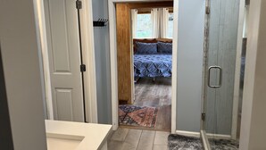 Uma casa de banho com chuveiro, lavatório e porta que dá para um quarto com cama e janela.