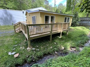 A small, single-story wooden cabin with a deck, surrounded by a grassy yard and a stream.