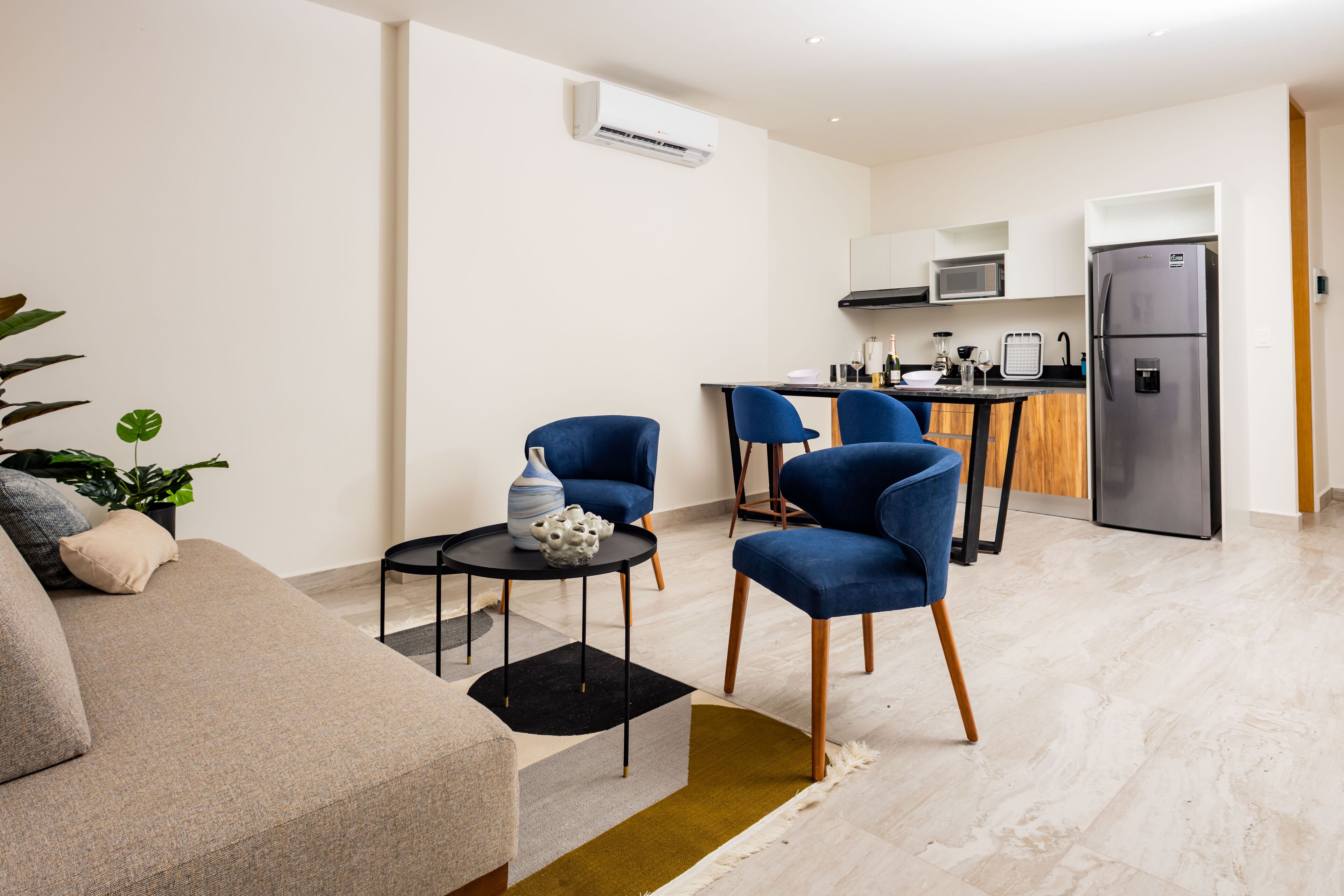 Una sala moderna con un sofá, dos sillones azules, una mesita con un florero, y una zona de cocina con nevera, microondas y fregadero.