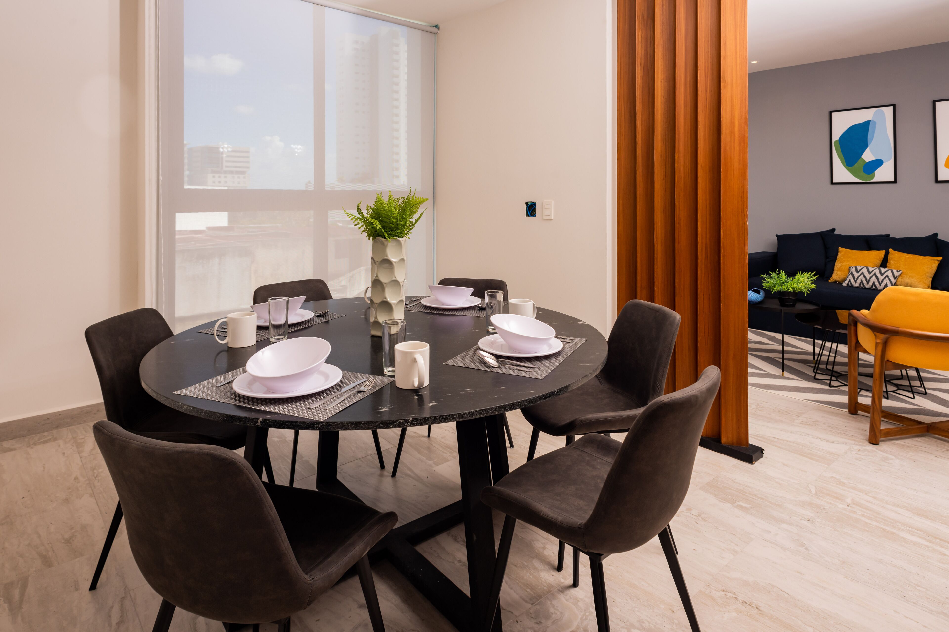 Un espace repas moderne avec une table ronde dressée pour six personnes, entourée de chaises foncées. On aperçoit un vase avec une plante sur la table, ainsi qu’une fenêtre donnant sur des bâtiments à l’extérieur.