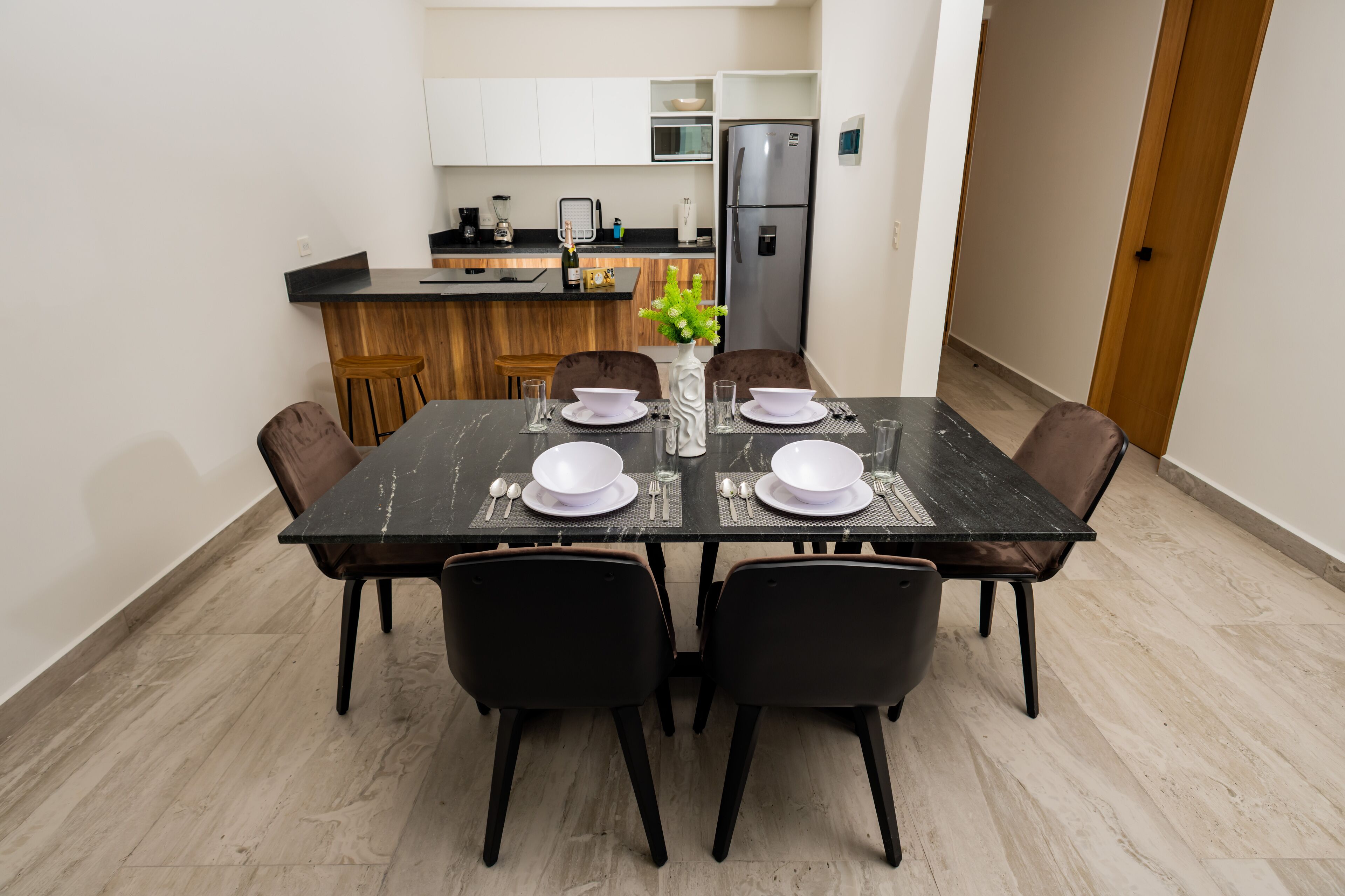 A dining area with a rectangular table set for four, surrounded by chairs. There is a kitchen in the background with white cabinets and a stainless steel refrigerator.