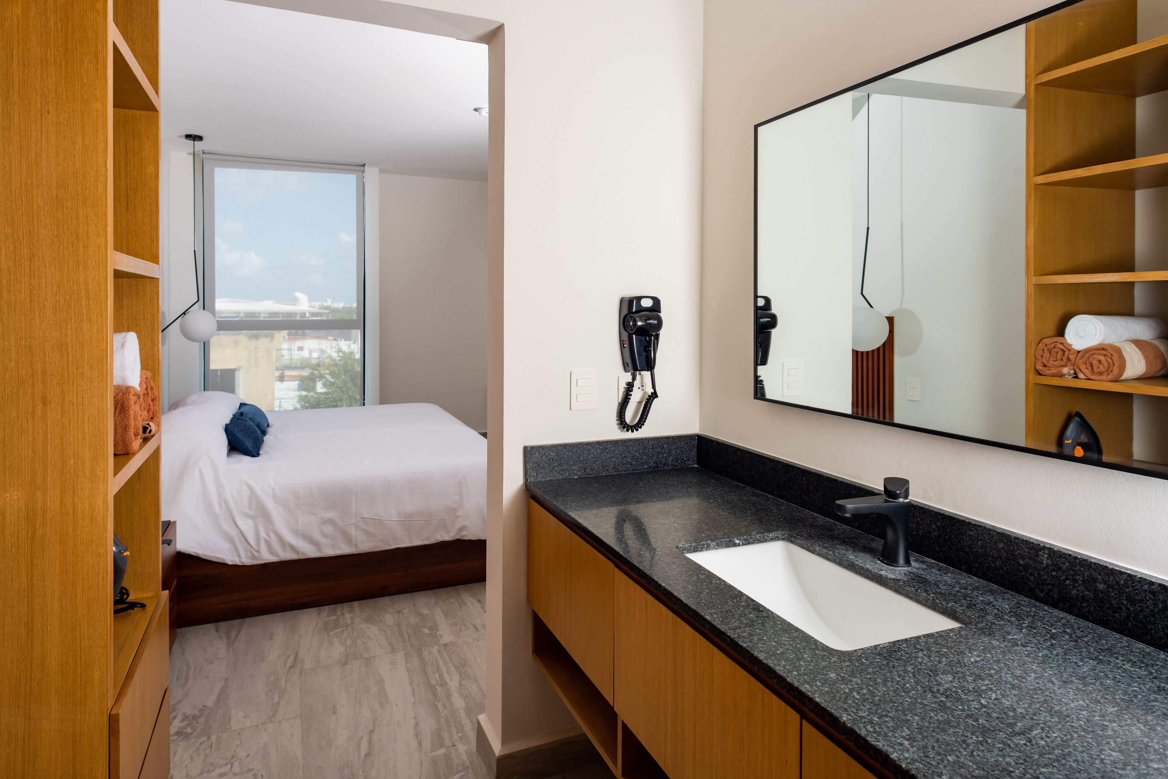 Una habitación de hotel moderna con una cama, un espejo grande, una encimera de granito negro y un tocador de madera.