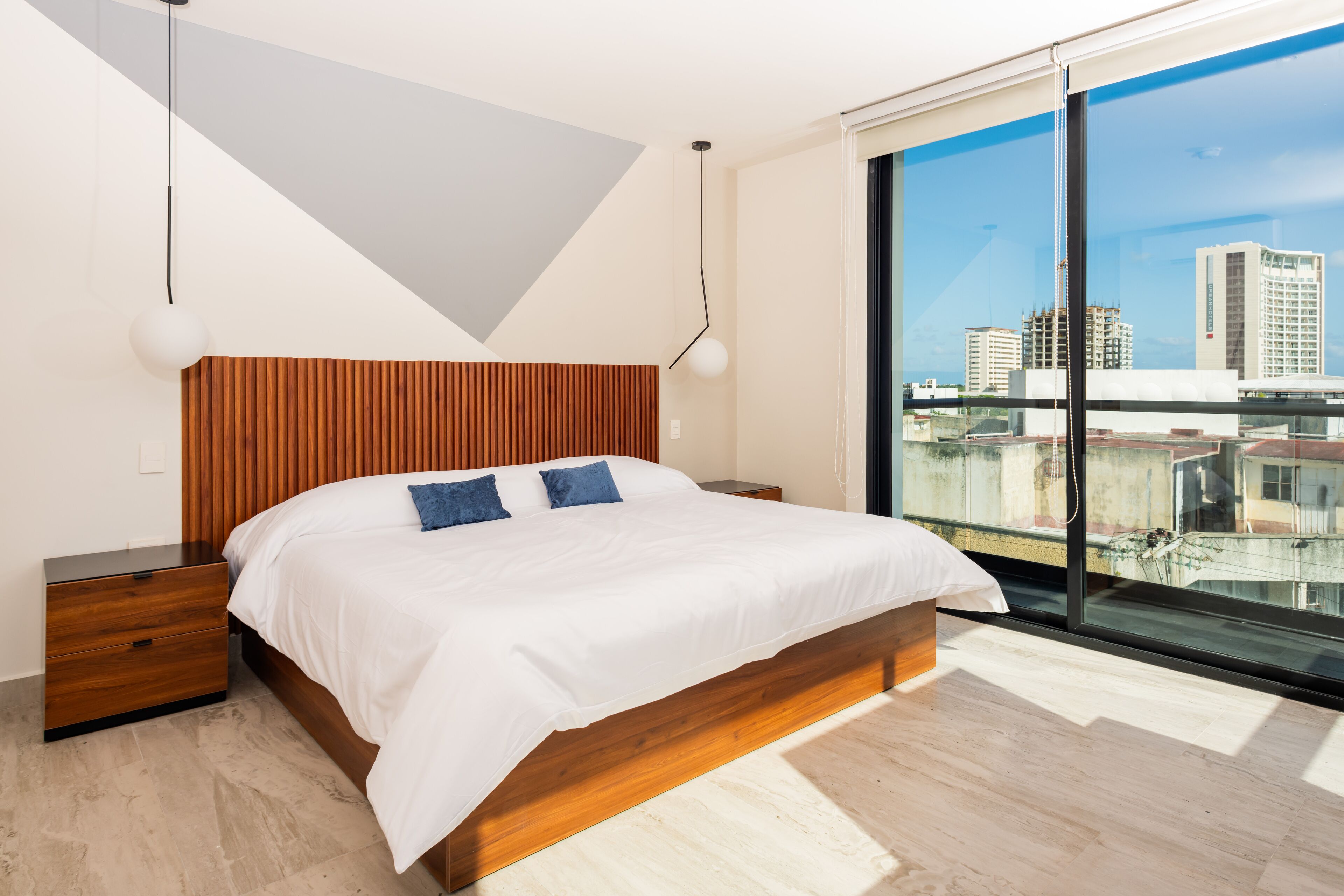 Une chambre moderne avec une tête de lit en bois, un grand lit avec une literie blanche, une table de chevet et une grande fenêtre donnant sur la ville.