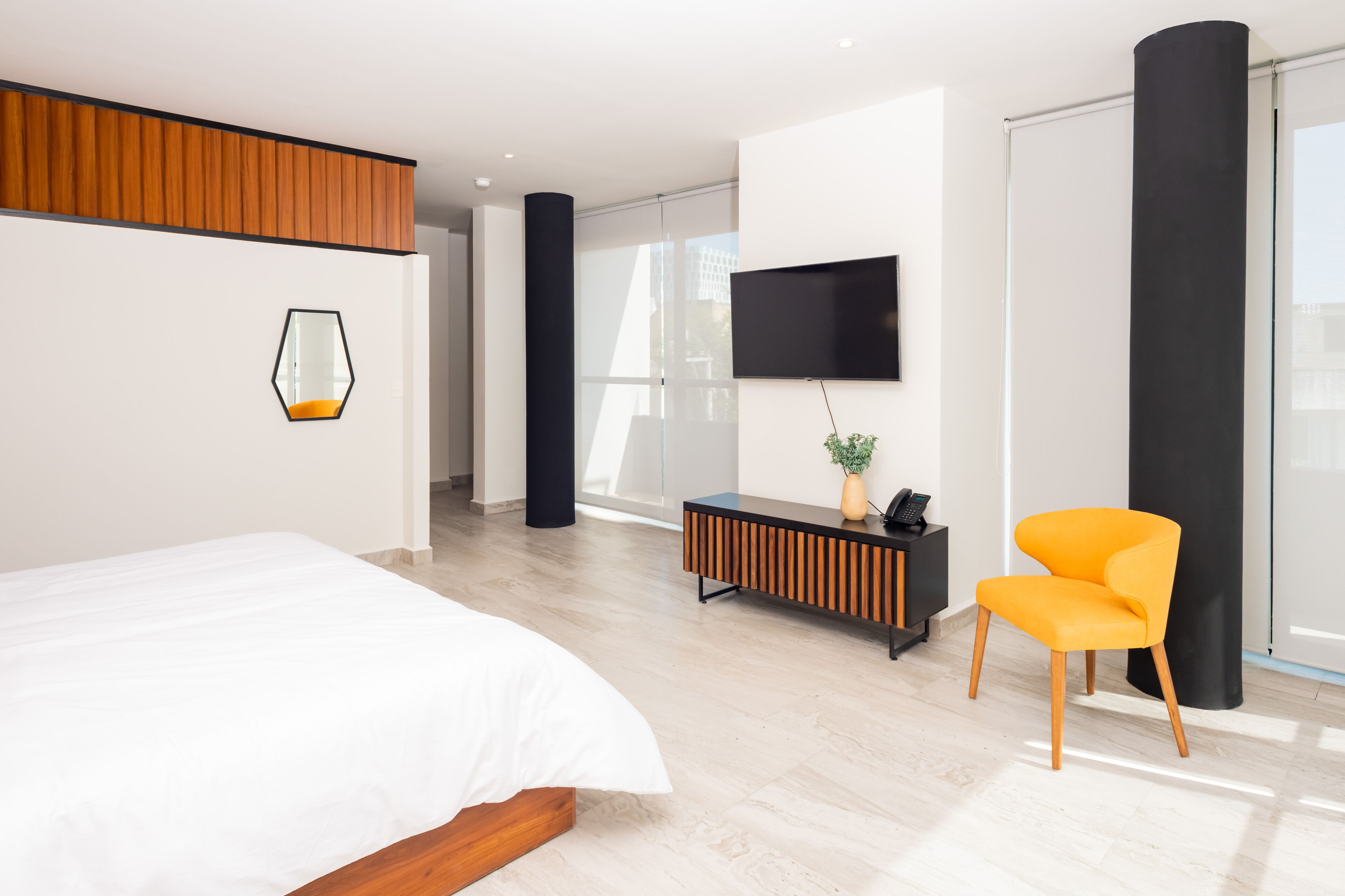 Una habitación de hotel moderna con una cama grande, un televisor montado en la pared, un panel de madera sobre la cama, un pilar negro y una silla de color naranja brillante.