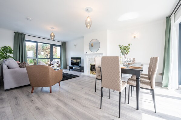 Un salon moderne comprenant un canapé, un fauteuil, une table à manger et des chaises. On y trouve également une cheminée, un téléviseur et une grande fenêtre avec des rideaux.