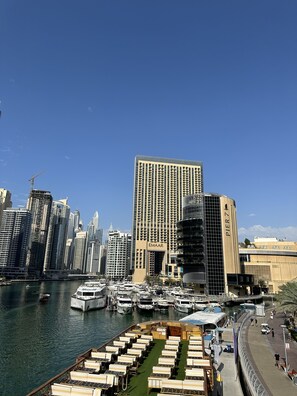 Uma marina com barcos atracados, um edifício grande com a inscrição 'EMAAR' e um calçadão com áreas de estar.